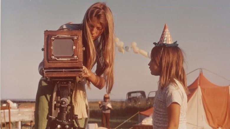An aged colour image of a young woman with long straight hair staring intensely at a large wooden box-type camera on a tripod. A young girl with medium straight brown hair watches intently while wearing a colourful birthday hat. There is a tent set up in the background.