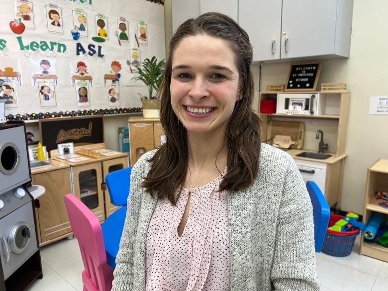 A woman in light pink top and a grey shrug smiles. She is in a preschool classroom.
