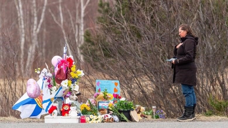 A woman in a black jacket places her hand on her heart as she looks at a roadside memorial of flowers, balloons and stuffed animals