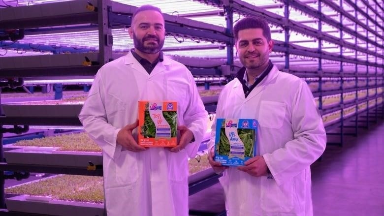 Two smiling men in white coveralls hold up green products in a warehouse featuring large trays of seeds.