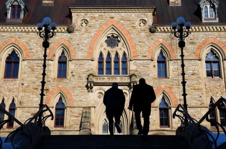 Two people silhouetted by the sun as they climb stairs toward a large stone building.