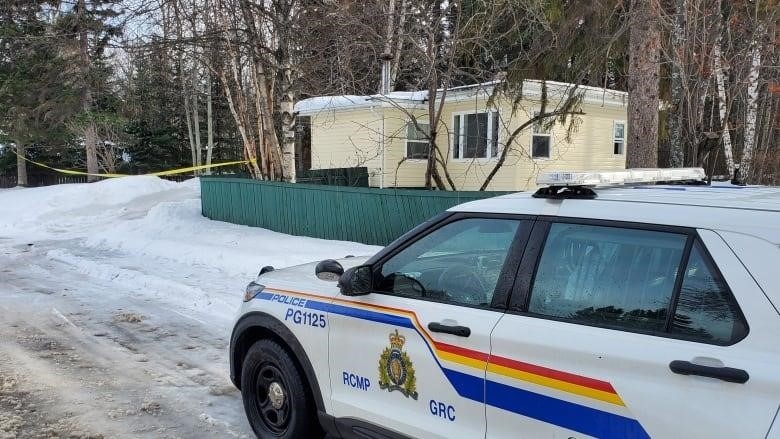 An RCMP cruiser sits on a snowy road leading to a yellow house surrounded by trees.