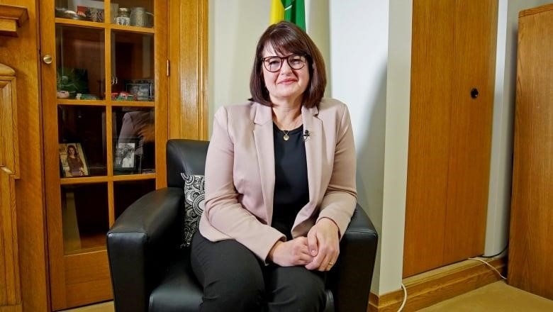 A woman in glasses sits in a black arm chair in front of the Saskatchewan flag. 