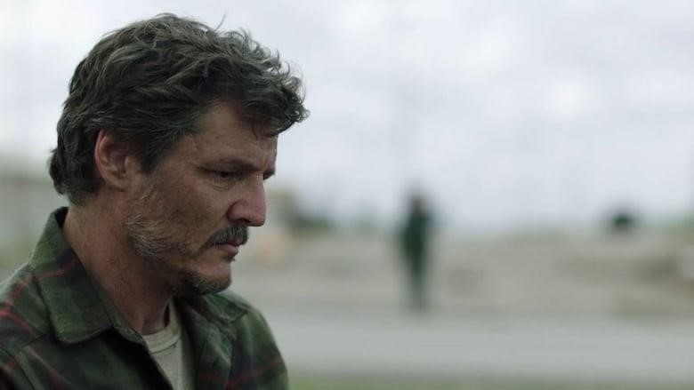 Still frame from the TV series The Last Of Us. Closeup shot of Pedro Pascal in the bottom left corner of the frame, wearing a green plaid shirt and looking down solemnly.