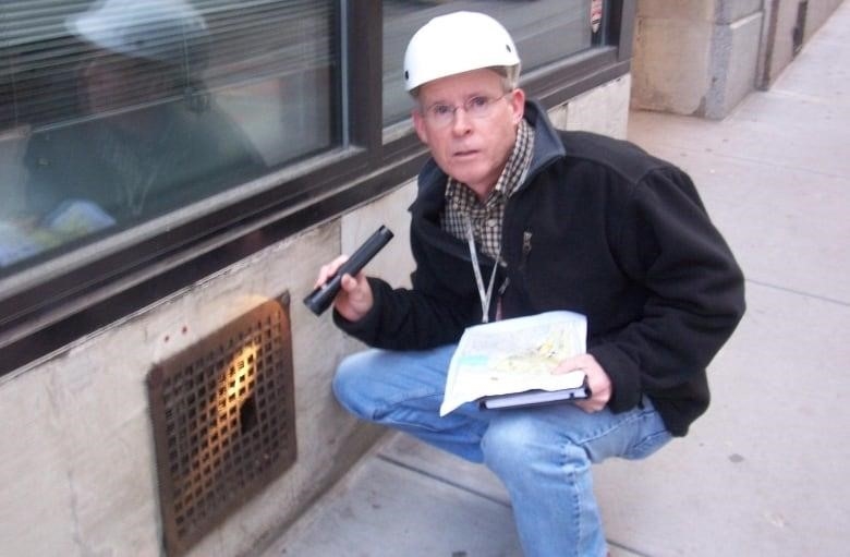 A man in a hardhat crouches on a side walk near a grate. 