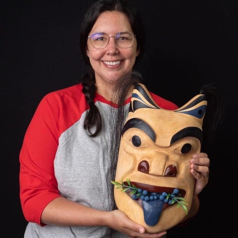 A woman stands with a wood carved mask in the style of the northwest coast First Nations. 