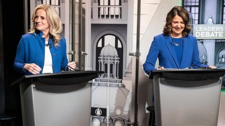 Rachel Notley looks off and to the right, while Danielle Smith checks her notes, both standing behind lecterns.