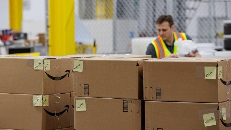 A row of cardboard boxes with Amazon branding is in the foreground, while a man wearing a high-visibility safety vest works in the background.