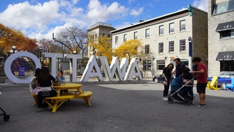 People sit at a picnic table or stand in front of a large white sign saying OTTAWA in a median.