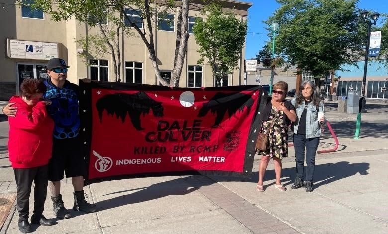People with sad faces hold up a red and black banner that says "Indigenous Lives Matter" and "Dale Culver Killed by Police" beneath the images of two bears and a full moon. 