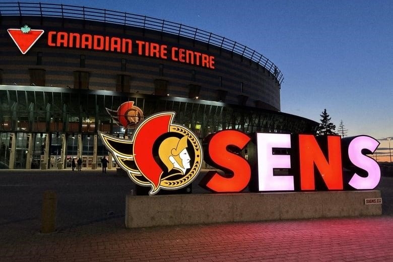 A red and white lit-up SENS sign in front of a hockey arena.