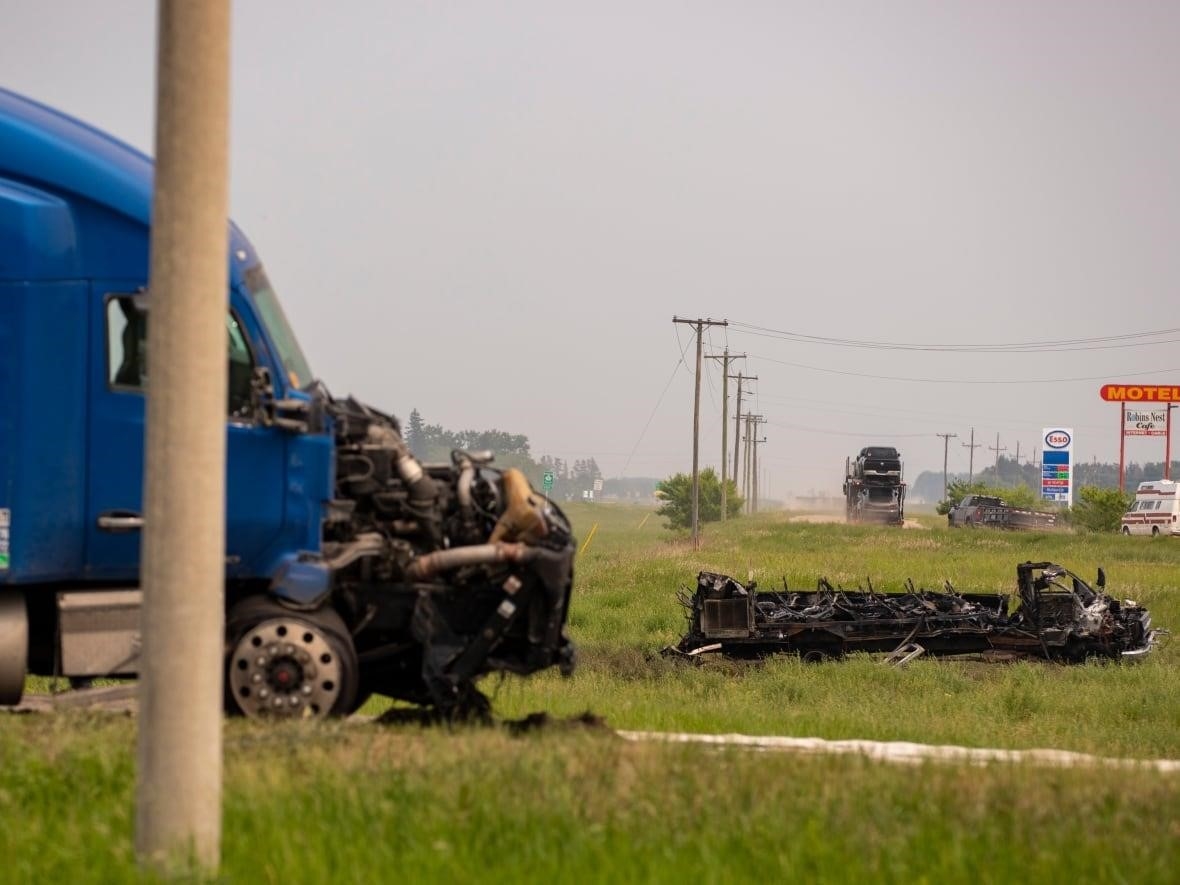 A semi trailer with a burned front end is pictured. A blackened passenger bus, destroyed by fire, is seen in the grass in the distance.
