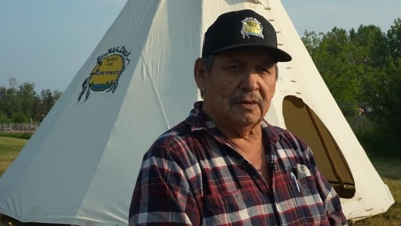 A man stands in the sun in front of a teepee