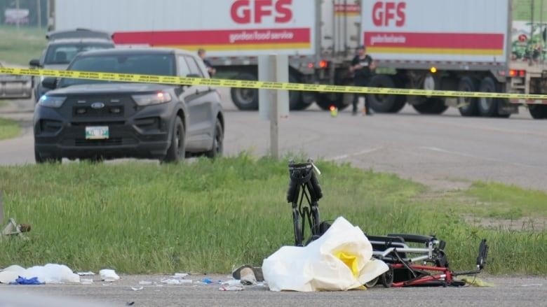 Two white and red semi-trailers are parked in the background while yellow police tape crosses the centre of the photo. A pair of folding walkers lie near the edge of a road. 