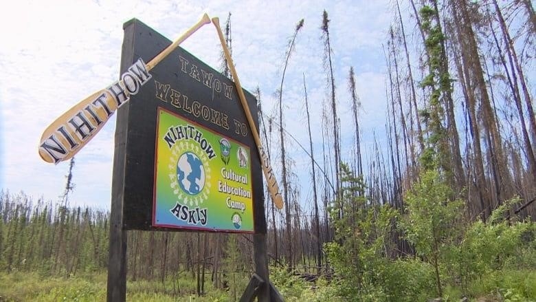 A colourful sign reading 'Nihithow Askiy Cultural Education Camp.' The forest on one side is lush and green, while the trees on the other side are charred and dead.
