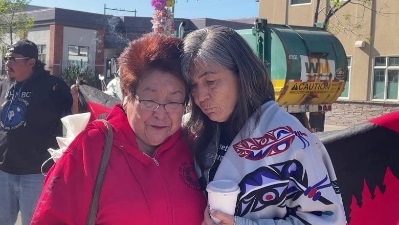 Two women lean against each other for support, both with somber faces. 