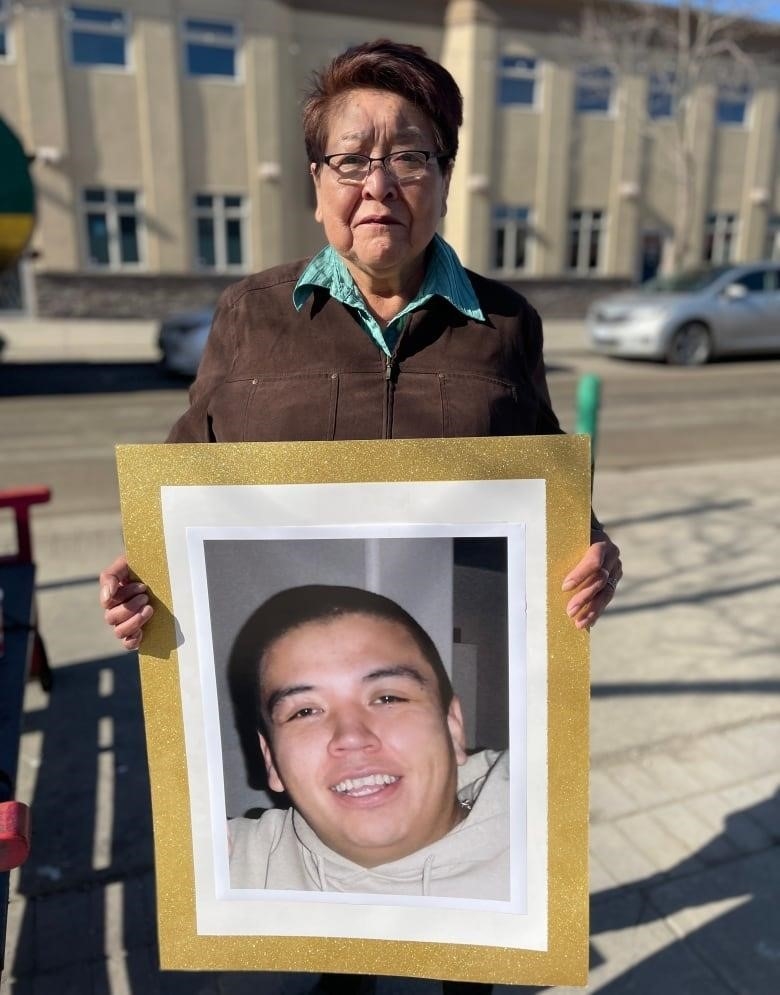 An Indigenous woman holds a framed picture of a smiling man outside a building.