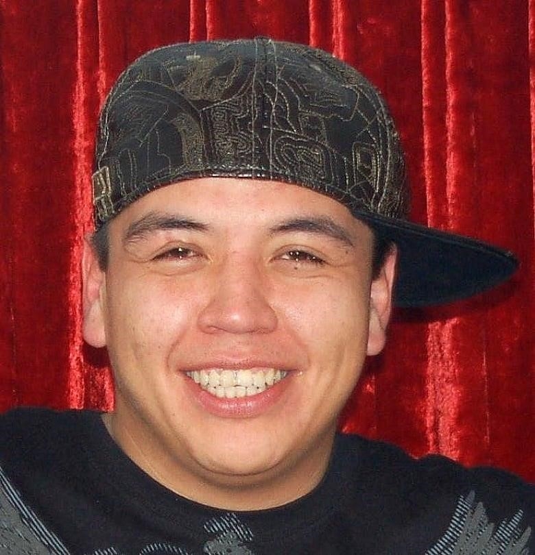 A youthful-looking Indigenous man with a sideways baseball cap smiles wide at the camera in front of a red velour background.