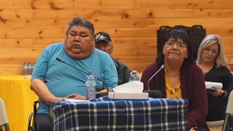 A man in a blue shirt sits in a wheelchair in front of a table with a microphone. A woman sits beside him. 