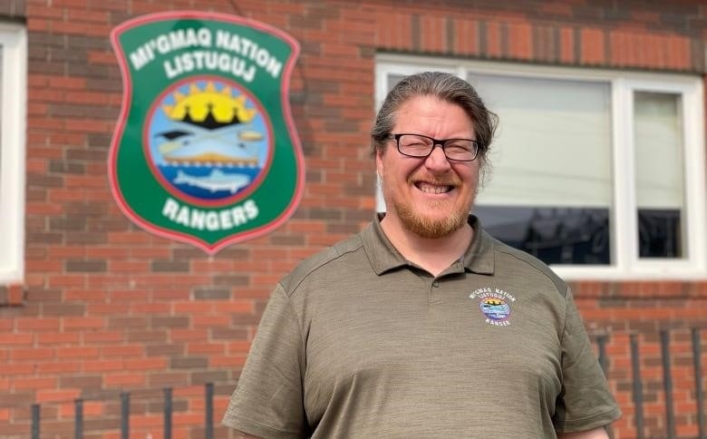 A man smiling outside a brick building with a green crest on it that says "MI'GMAQ NATION LISTUGUJ RANGERS"