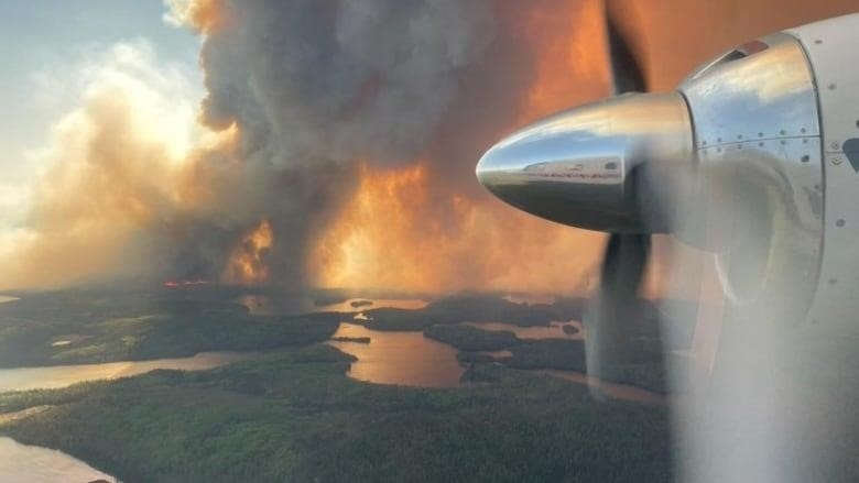 An airplane flies past a large wildfire burning amongst forests and lakes