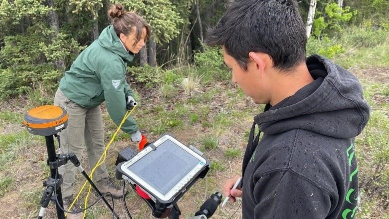 A woman in a green jacket and a man in a black hoodie adjust a screen on a piece of radar machinery while standing in a patch of grass near a wooded area.