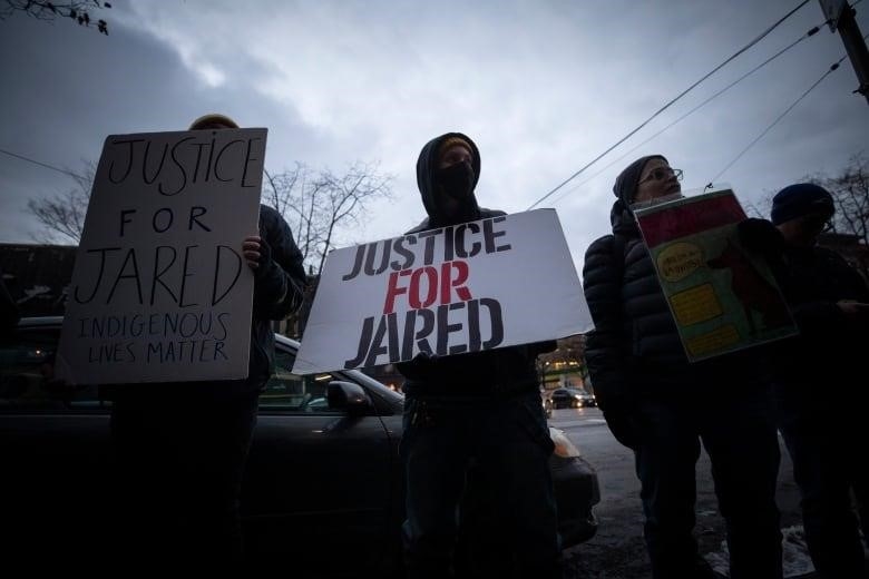 A person at a rally holds a hand-made sign that says "Justice for Jared" beneath a grey sky. 