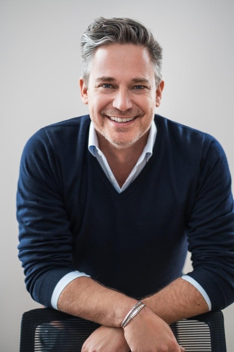 A smiling man standing and resting his arms on a chair.