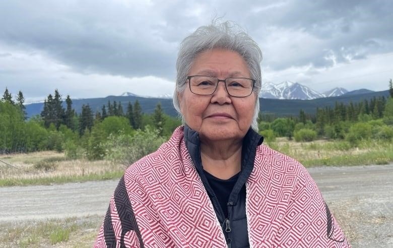 An Indigenous woman with short grey hair and glasses, wrapped in a red, white and black blanket, poses for a portrait outdoors under a wide, cloudy sky.