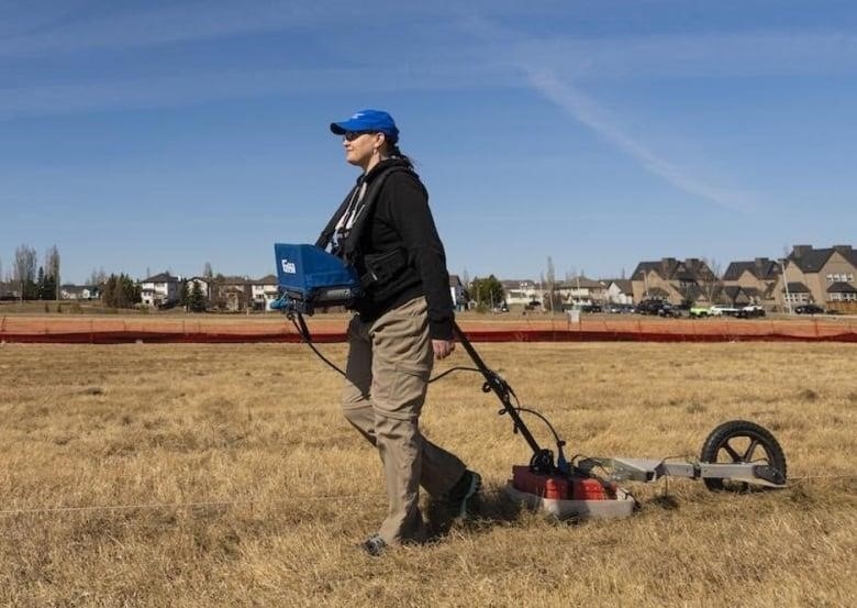 A woman in a blue baseball cap walks across a field. She pulls a machine behind her along the grass.