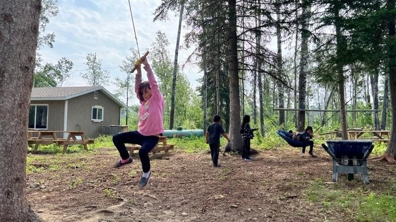 A young student in a pink sweater holds onto a piece of wood attached to a tall tree. She's swinging around the trunk while her classmates play behind her.