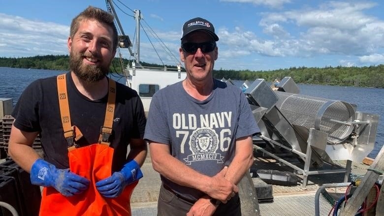 Two people stand next to each other on a boat. Person on the left is wearing orange overalls and gloves. The person on the right is wearing an Old Navy t-shirt and a ball cap.