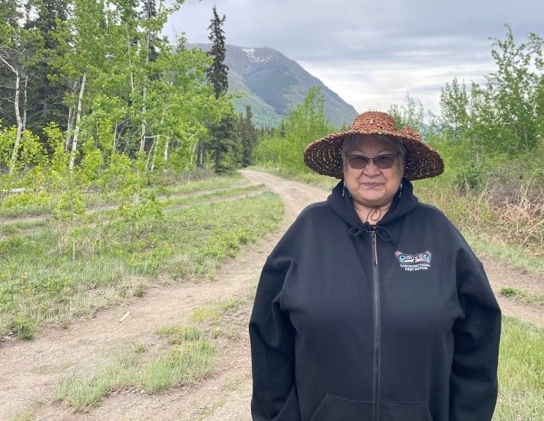 An Indigenous woman, wearing a wide-brimmed straw hat, sunglasses and a black jacket, poses for a portrait outdoors, standing along a dirt road that cuts through a grove of trees.