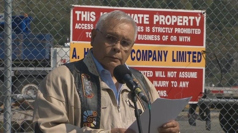 A man wearing a traditional vest with colourful patches and a tan-coloured shirt holds a piece of paper and speaks into a microphone.