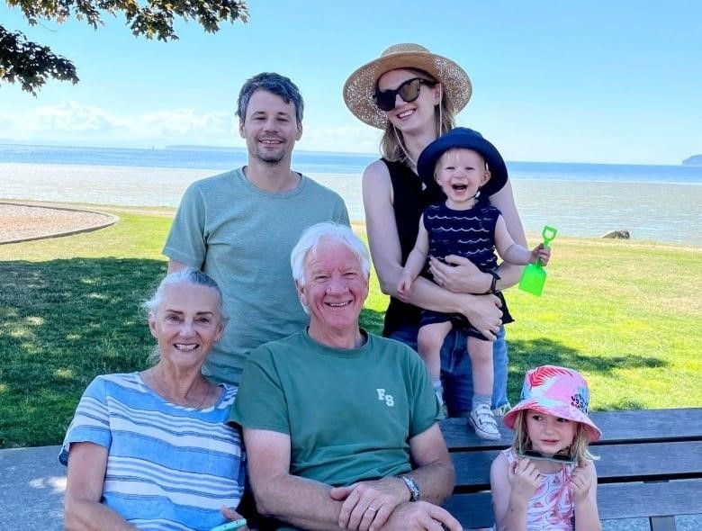 A family of six smiles for the camera, including two young children, two parents and two grandparents sitting on a park bench by a large body of water.