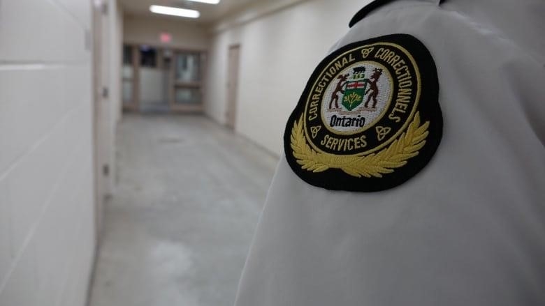 A shoulder patch reading Ontario Correctional Services is seen on someone wearing a white shirt in a jail corridor.