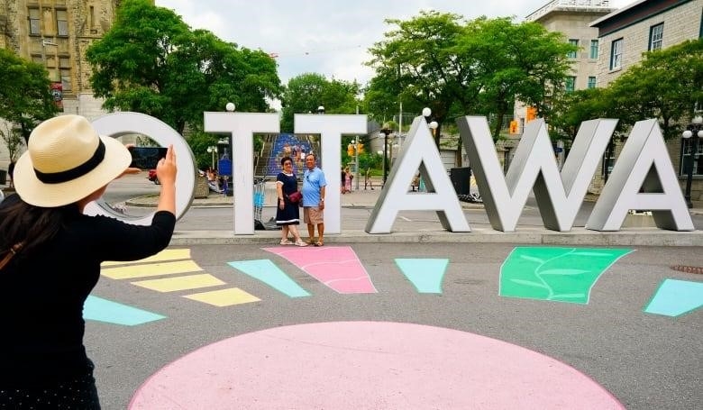 A person takes a photo of two people standing in front of a large "Ottawa" sign.