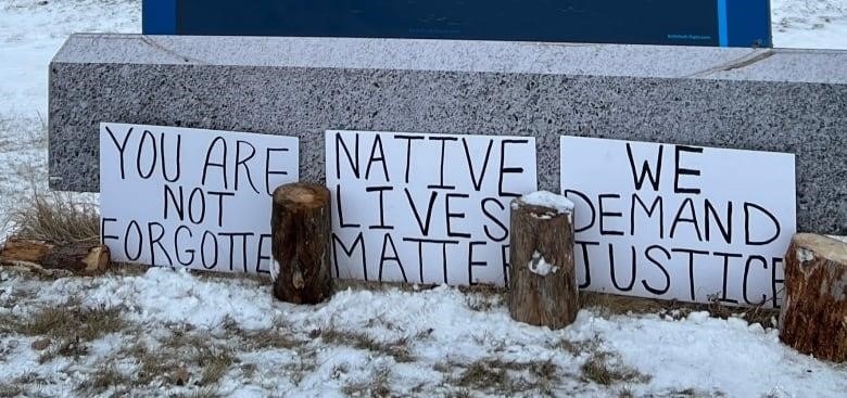 Handwritten signs reading "You are not forgotten", "Native lives matter" and "We demand justice" are leaned up against the concrete base of a larger sign.