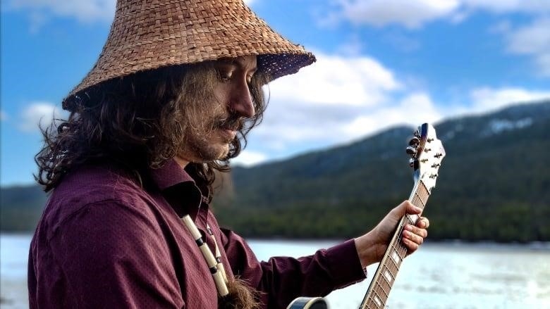 A man stands by the mouth of a river wearing a cedar hat, holding an electric guitar.