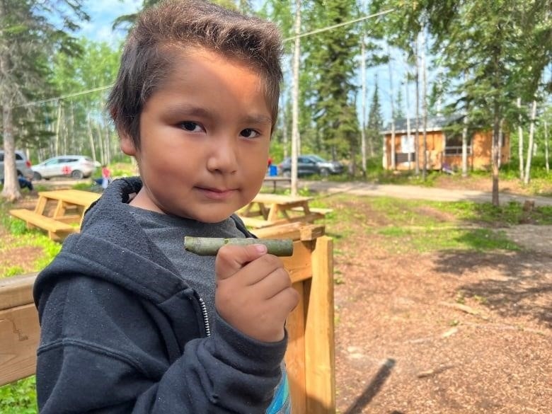 A young boy holds up a whistle made of a poplar tree branch. Behind him, you can see tall trees and a cabin.