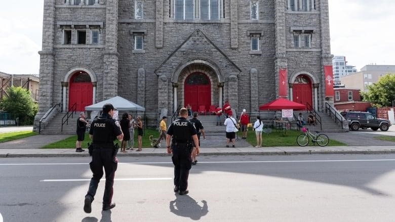 Police officers walk across a street, heading toward several people standing in front of a church building with bright red doors.