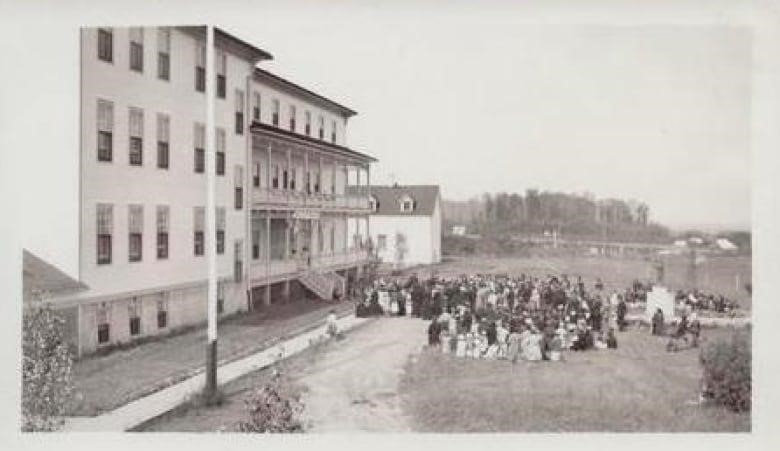 A black and white photograph, shot from afar, shows a crowd of young people ion the grass near the steps of a large complex of buildings. 