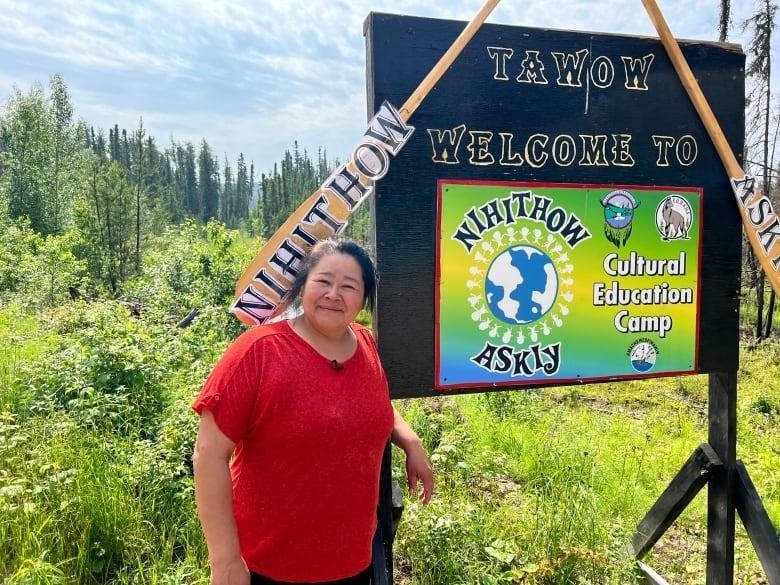 A smiling woman wearing a red shirt stands next to a colourful sign. The sign welcomes people to the land-based cultural education camp near Stanley Mission in northern Saskatchewan.