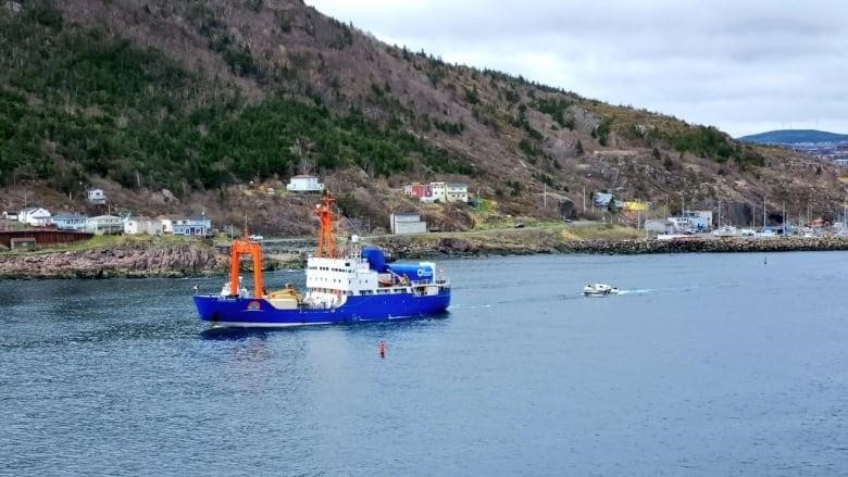 A large blue ship is seen pulling a large, white submersible, floating on the water, in a wide shot of the St. John's harbour. 