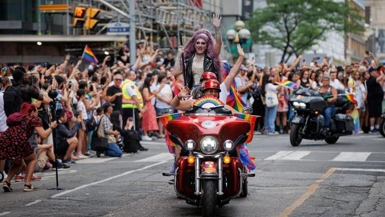Three people, including a person in drag, wave to the crowd while on a motorcycle that's part of a Pride parade.