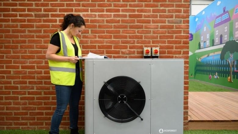 A woman in a safety vest looks at some papers resting on the outdoor unit of a heat pump
