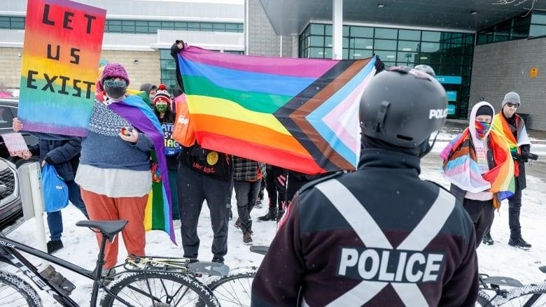 People with rainbow flags stand in front of a building as a police officer looks on. 