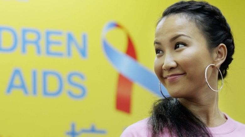 A woman with braised hair, earrings and in a pink top looks to her right in front of a yellow wall with a ribbon at the launch of a campaign in Hong Kong focused on the enormous impact of HIV/AIDS on children.