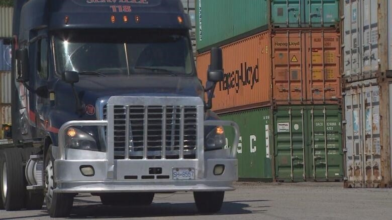 The front end of a dark coloured transport truck is shown in front of rows of shipping containers.