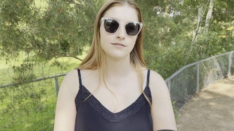 A young woman wearing a hat and sunglasses looks at the camera while standing outside.